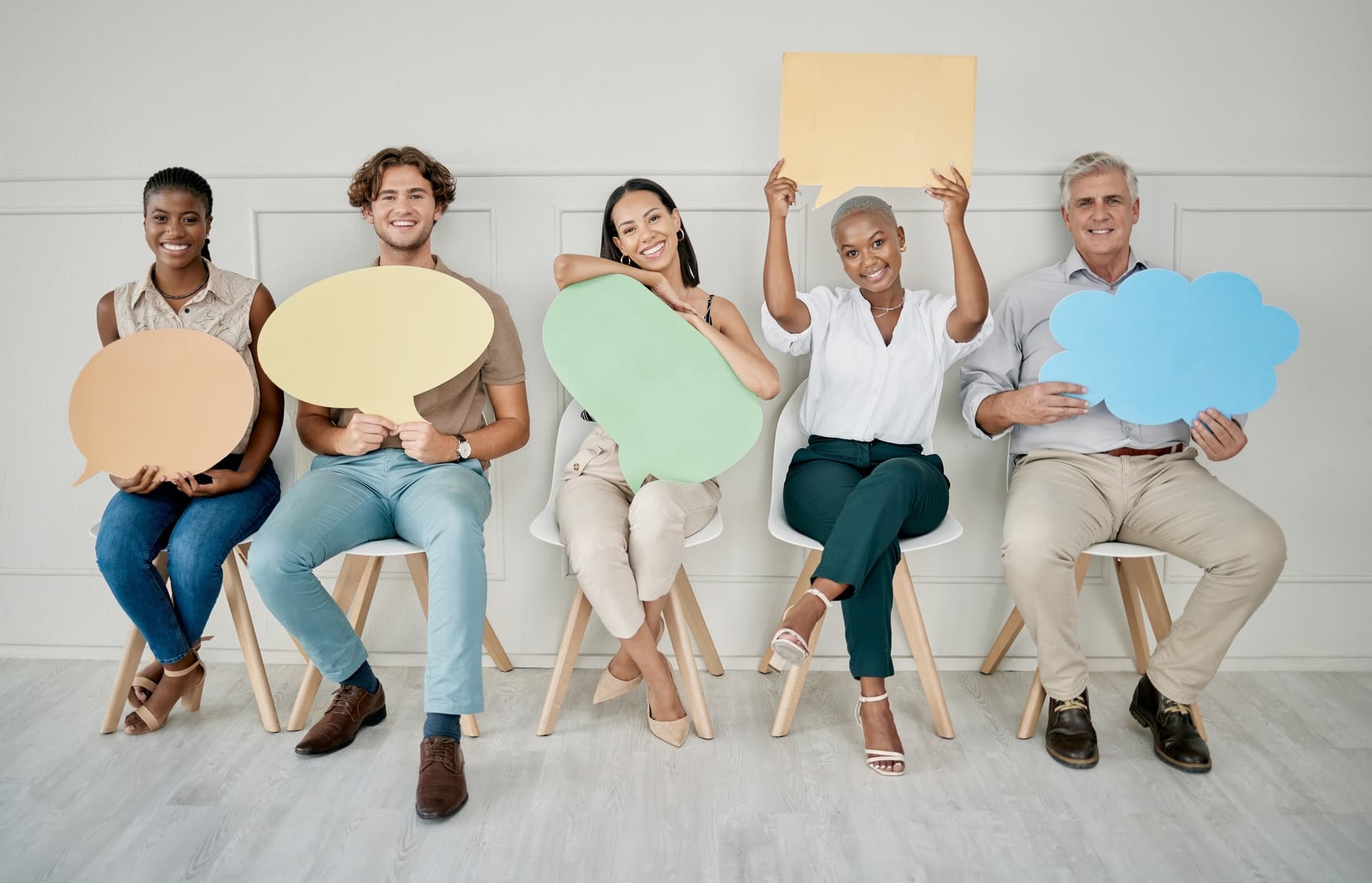 Five people on white chairs, each holding a pastel speech-bubble cutout.