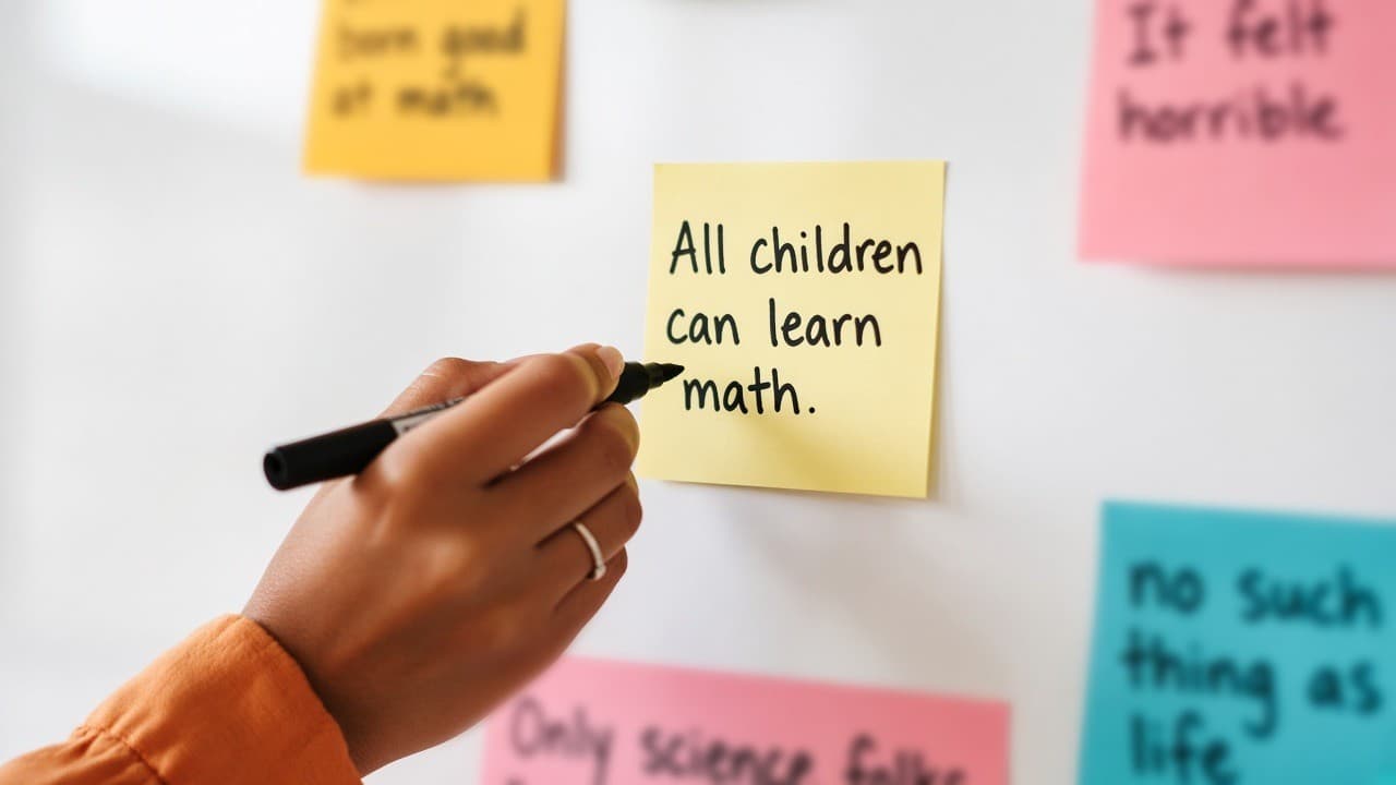 A hand writing “All children can learn math.” on a yellow sticky note, surrounded by colorful notes on a whiteboard.